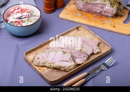 Roasted turkey thigh on a wooden board, cut into pieces. In the background is a bowl of vegetable salad and a turkey. Close-up. Stock Photo
