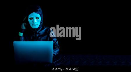 Hacker in mask and hoodie using a laptop to browse the net and serious face thinking about question Stock Photo