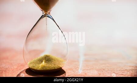 Gold Sand running through the bulbs of an hourglass measuring the passing time in a countdown to a deadline, on a light pastel reddish background with Stock Photo