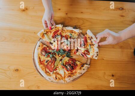 Overhead shot of two female hands taking slices from the whole pizza Stock Photo
