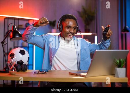 Overjoyed young man in headset screaming and gesturing while celebrating victory in video games. African american guy using wireless laptop and joystick during leisure time. Stock Photo