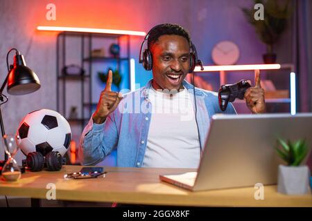 Emotional black man in headset looking at computer screen and feeling satisfaction during victory in video games. Happy guy playing games with joystick alone at home. Stock Photo