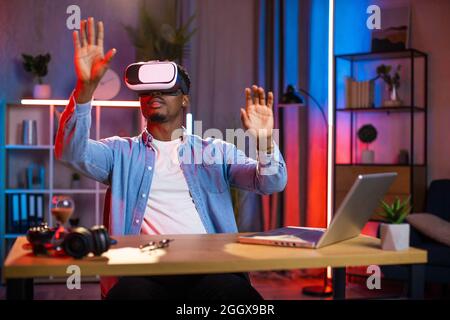 African american man sitting at home office and using VR glasses and laptop for remote work. Black male freelancer touching virtual screen with fingers. Stock Photo