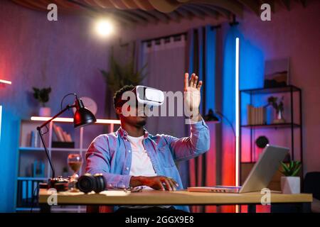 African american man sitting at home office and using VR glasses and laptop for remote work. Black male freelancer touching virtual screen with fingers. Stock Photo