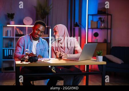 Two multi ethnic coworkers smiling and talking while sitting at workplace during evening time. Arabian woman in hijab and black man in denim shirt using laptop for remote work. Stock Photo