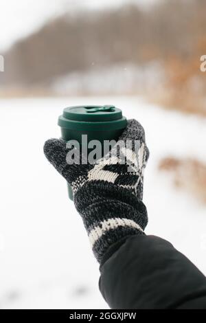 Hand in cozy glove holding reusable cup of tea or coffee on background of snowy lake in winter. Hiking and traveling in cold winter season. Warm drink Stock Photo