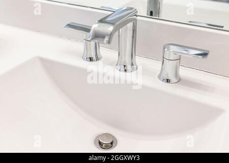 New modern stainless steel metal faucet with handles in bathroom room white porcelain sink closeup by mirror with drain hole Stock Photo