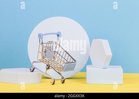 Mini shopping trolley cart with geometric podium platforms on yellow and light blue background. Online shopping concept. Front view Stock Photo
