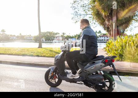 Miami Beach, USA - January 19, 2021: Young man riding motorcycle scooter in danger of accident while using smartphone mobile phone on city street road Stock Photo