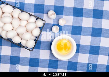 A small crate of fresh eggs with two cracked eggs rests atop a tabletop covered in a traditional country blue and white checkered tablecloth. Stock Photo