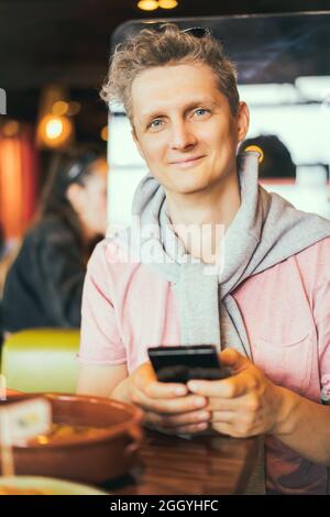 Portrait of a smiling young man using mobile phone while sitting in cafe. Make food photo for social networks, blogging, chatting, watching the video Stock Photo