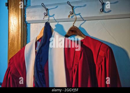 Two red gowns or traditional pastor robes hung on vintage wooden hangers. One long robe has a wide white collar. The cassock is for ministers to wear. Stock Photo