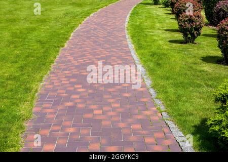 curved stone tile pavement crescent, path in park landscaped with green grass and bushes, garden landscape on a sunny summer day. Stock Photo