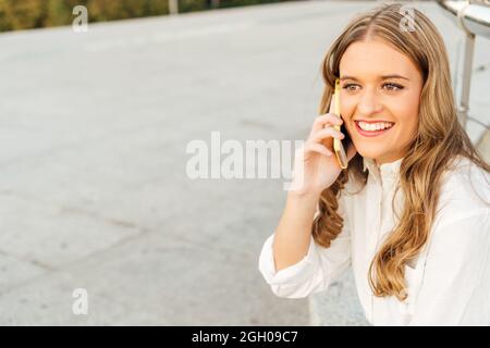 Portrait of a young blonde woman with long hair and white shirt talking on the smart phone on the city street. Stock Photo