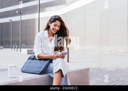 horizontal portrait of an elegant young indian woman. She is sitting near office building and using her smart phone Stock Photo