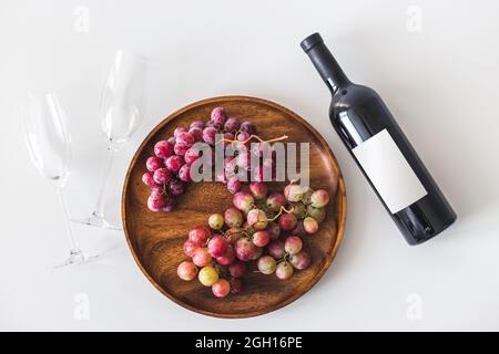 Red wine bottle, large burgundy fresh grapes on round wooden dish, empty wine glasses on white background, copy space flat lay, top view, mockup.