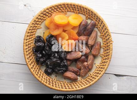 Several appetizing dates, dried apricots, prunes in a ceramic plate on a wooden table, close-up, top view. Stock Photo