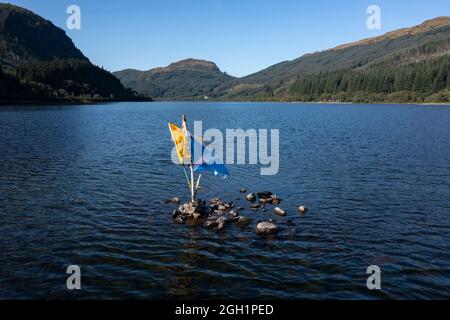 Loch Lubnaig, Callander, Loch lomond and Trossachs National Park, Scotland. 2 September 2021. PICTURED: The Saltire & Lion Rampant (Royal Banner) flags poke out of the water in the middle of Loch Lubnaig. Loch Lubnaig (Loch Lùbnaig in Gaelic) is a small freshwater loch near Callander in the Stirling council area, Scottish Highlands.  The loch nestles in the space between Ben Ledi and Ben Vorlich. Fed by the River Balvaig from the north and drained by the Garbh Uisge to the south, Loch Lubnaig offers fishing from the shore while canoes can be rented at the north end. Alternatively, two car park Stock Photo