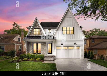 A new, white modern farmhouse with a dark roof and black windows. The left side of the house has a light rock siding and covered front porch at sunset Stock Photo