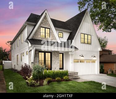 A new, white modern farmhouse with a dark roof and black windows. The left side of the house has a light rock siding and covered front porch at sunset Stock Photo