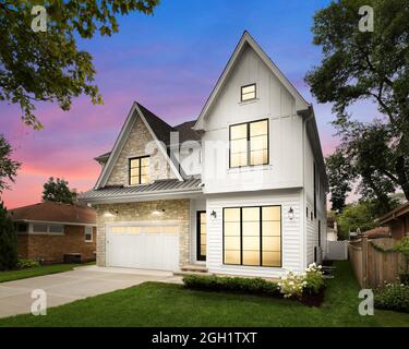 A new, white modern farmhouse with a dark shingled roof and black windows. The left side of the house is covered in a rock siding at sunset. Stock Photo