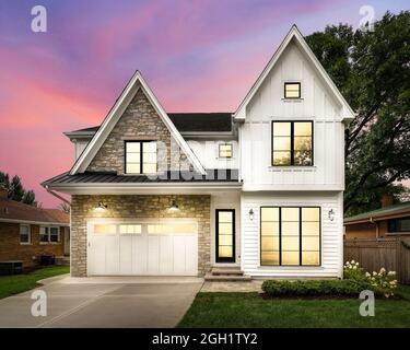 A new, white modern farmhouse with a dark shingled roof and black windows. The left side of the house is covered in a rock siding at sunset. Stock Photo