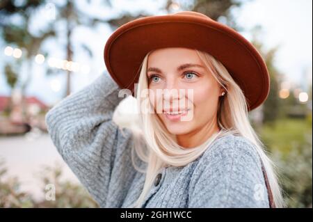 Beautiful blonde smiling adult girl 20-24 year old wear casual warm clothes and hat posing in city park. Autumn season. Romantic lady outdoors. Stock Photo