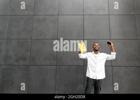Overjoyed african american businessman or student looking up, excited about good news. Stylish male employee holding yellow laptop and smartphone Stock Photo