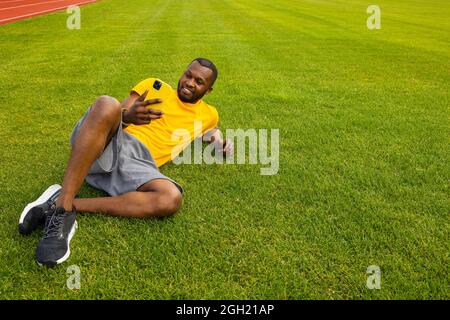 Joyful handsome african american male laying on the grass, relaxing and taking a break after workout. Smiling happy man using smartphone, talking with Stock Photo