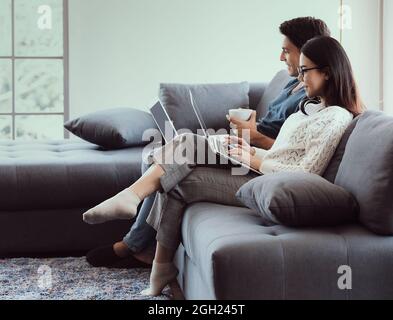 Mix race family lovers, Caucasian husband and Asian wife, sitting in livingroom working via laptop computer on sofa in easy relax gestures. Concept of Stock Photo