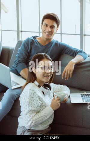 Mix race family lovers, Caucasian husband and Asian wife, sitting in livingroom working via laptop computer on sofa in easy relax gestures. Concept of Stock Photo