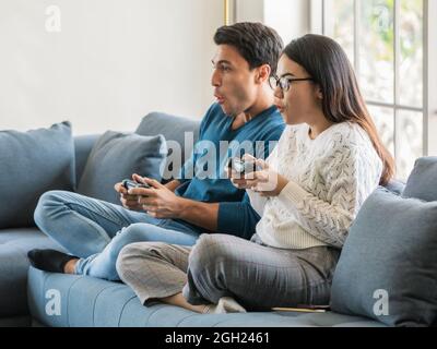 Handsome Caucasian man and beautiful Asian woman lovers sitting in house living room and using joystick to play game togehter with excited faces. Dive Stock Photo