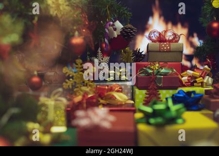 Pile of shiny luxury colorful gift boxes. Christmas and new year celebrate gifts stack beside full decorated Xmas tree with flame of fire in fireplace Stock Photo