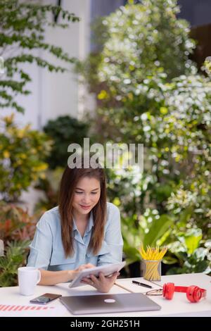 Businesswoman sitting at working desk using tablet and laptop computer connect to internet in small garden green background corner at home. Concept of Stock Photo