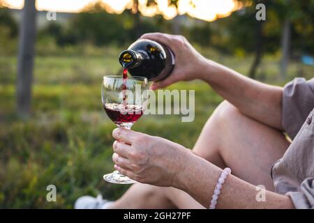 Woman sitting in vineyard and pouring red wine into drinking glass. Relaxation with alcohol drink during sunset outdoors Stock Photo
