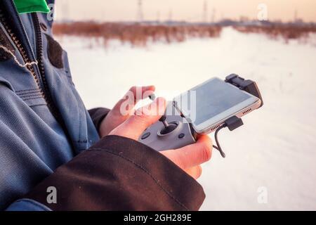 Close up view of hands of a man holding wireless remote control of drone with phone wintertime in the field. Stock Photo