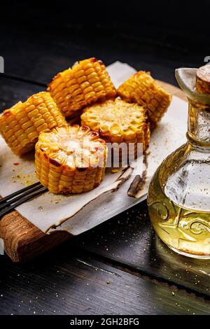Close-up grilled sweet corn on the cob with oil and salt on a wooden board, close-up on a dark wooden background. Delicious grilled corn and a bottle Stock Photo