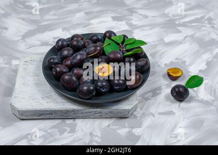 Fresh ripe plum fruits whole and chopped in a bowl and on a wooden cutting board, stone-concrete background. Close-up. Stock Photo
