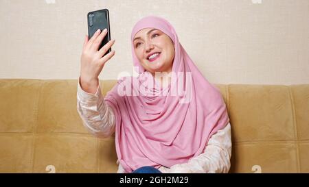 Beautiful muslim woman in hijab talking by video call while sitting on the couch Stock Photo