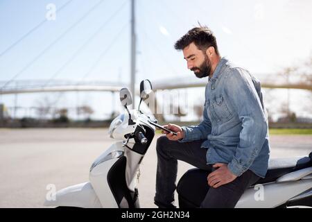 Young Spanish man using cell phone or smartphone sitting on his motor scooter outdoors in the city. Businessman checking work issues on cell phone. Stock Photo