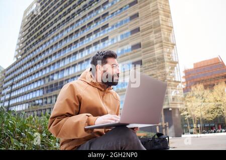 Young businessman works with laptop outdoors. Entrepreneur in casual clothes in a corporate area. Man working remotely on the street of buildings. Stock Photo