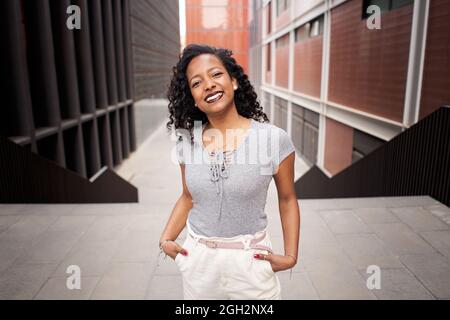 Attractive businesswoman on city street looking at camera outdoors near large office building. Casual dressed young woman with cheerful look, smiling Stock Photo