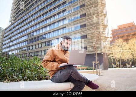 Business man typing on laptop computer outdoors. Casual dressed man working with laptop sitting on a bench. Concept of telecommuting. Remote work. Stock Photo