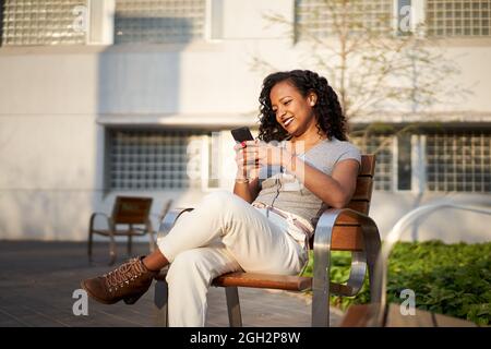 Young beautiful woman typing on cell phone sitting on bench- Smiling mixed race woman using smartphone, city at sunset.  Stock Photo
