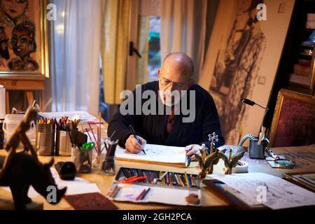 Artist working in the studio. Older man painting a picture using a magnifying glass in a creative and chaotic environment. Stock Photo