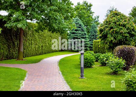 stone tile footpath curve arcing in the park among green plants of evergreen thuja hedges and trees with deciduous bushes and iron ground garden lante Stock Photo