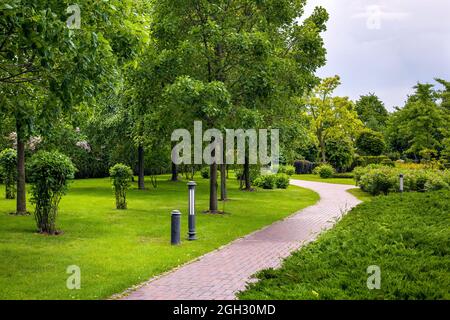 stone tile walkway curve arcing in the park among green plants of evergreen thuja hedges and trees with deciduous bushes and iron ground garden lanter Stock Photo