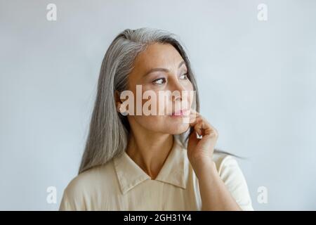 Thoughtful grey haired woman in stylish shirt looks aside on light background Stock Photo