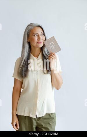 Thoughtful hoary haired Asian lady customer holds gift card standing on light grey background Stock Photo