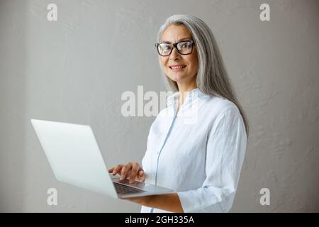 Positive hoary haired Asian woman in white blouse touches pad of laptop in studio Stock Photo